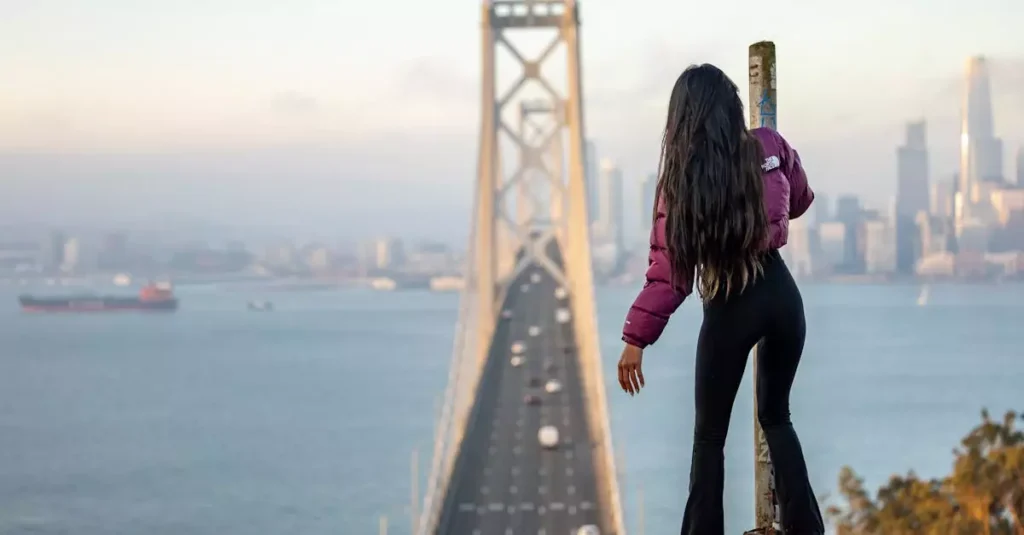 Best Adventure Bridge Climbs in the World 1 Woman standing on pole with a view of San Francisco Bay Bridge, capturing urban and adventurous spirit.