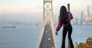 Woman standing on pole with a view of San Francisco Bay Bridge, capturing urban and adventurous spirit.