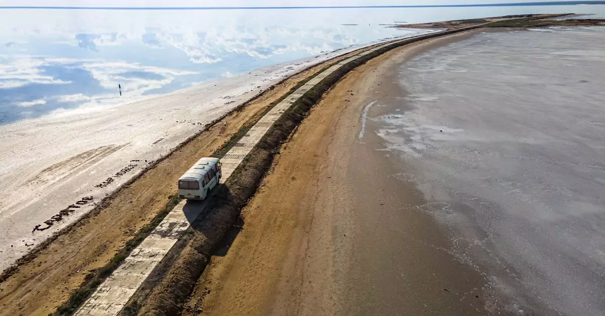 Aerial shot of a campervan traveling along a quiet coastal road with reflective seas and an empty beach.
