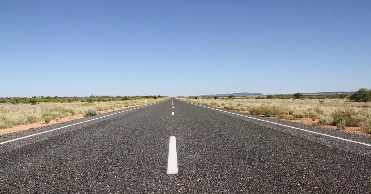 A long straight road stretches through the Australian outback under a clear blue sky.