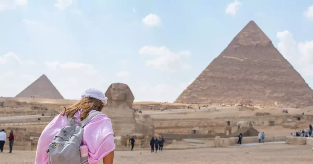 A tourist in a pink shirt wanders the Pyramids of Giza and Sphinx under a clear sky.
