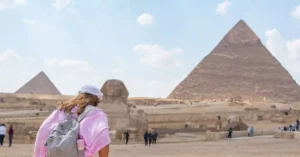 A tourist in a pink shirt wanders the Pyramids of Giza and Sphinx under a clear sky.
