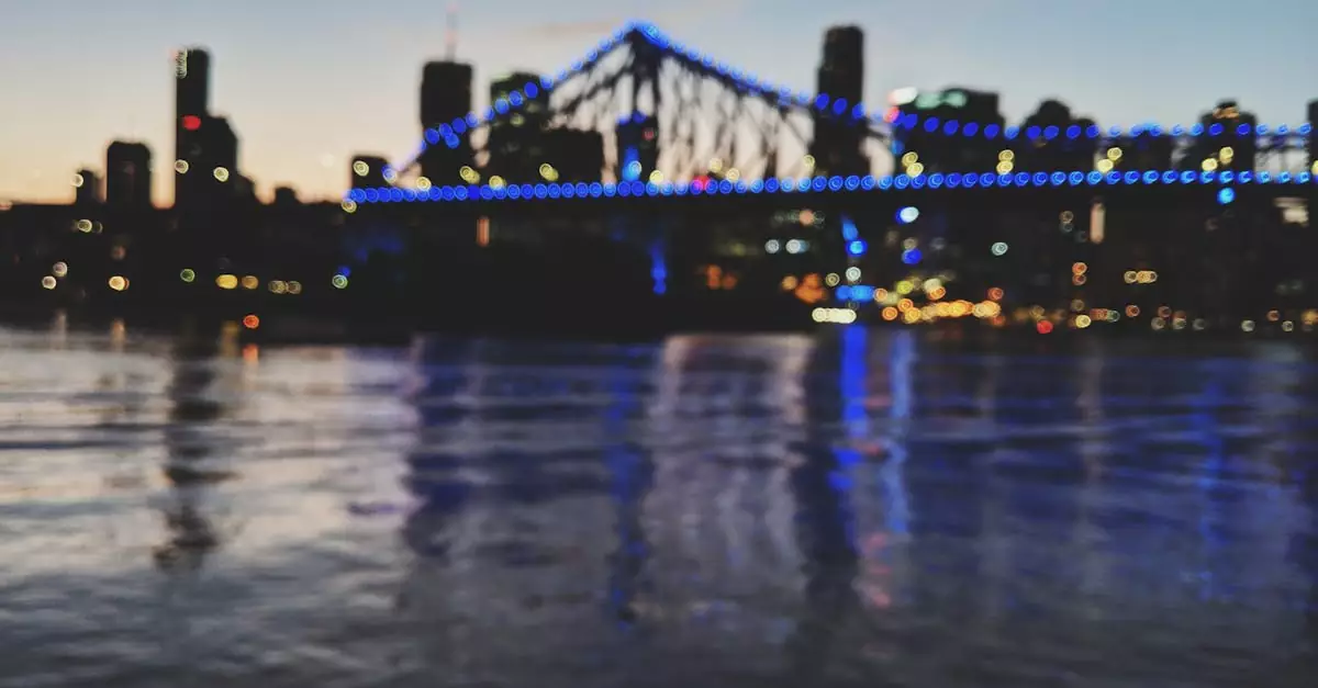 A blurred night view of the illuminated Story Bridge and Brisbane skyline reflecting on the river.