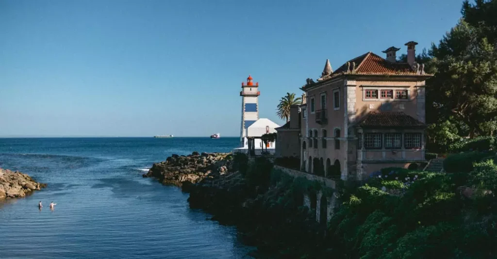 Scenic view of Santa Maria Lighthouse by the Cascais coast with a historic building and clear sky.