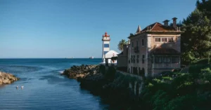 Scenic view of Santa Maria Lighthouse by the Cascais coast with a historic building and clear sky.