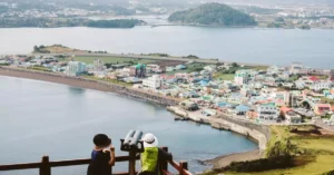 Tourists enjoying a panoramic view from Seongsan Ilchulbong overlooking Jeju Island and coastal village.