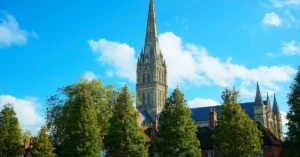 Elevated view of the iconic Salisbury Cathedral spire surrounded by lush trees under a bright blue sky.