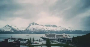 A cruise ship docked with snowy mountains in the background, under a cloudy sky.