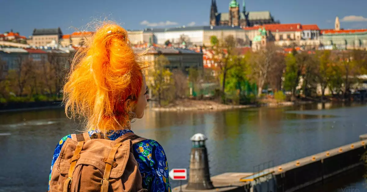 A vibrant-haired woman with a backpack gazes over Prague's scenic cityscape and river.