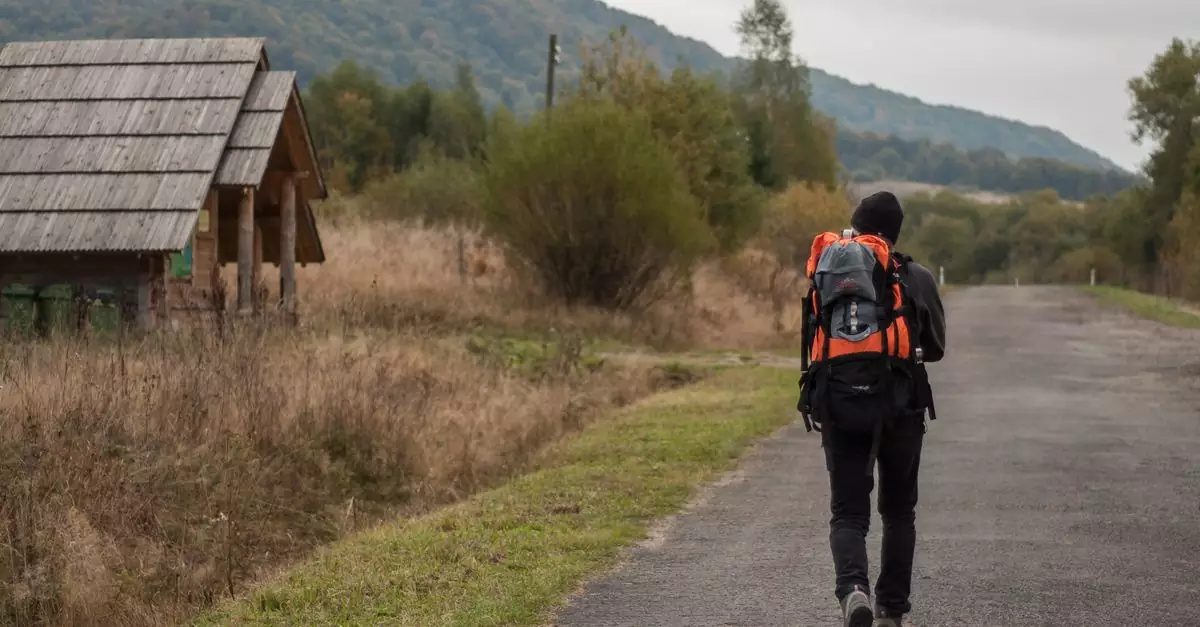Back view full body of anonymous hiker with backpack strolling on asphalt roadway near shabby house inn suburb area during trekking