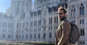 A man with a backpack stands in front of the historic Hungarian Parliament Building in Budapest.