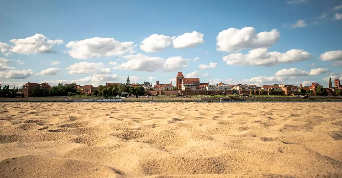 A scenic view of a sandy beach with a historic city skyline under a bright blue sky with clouds.