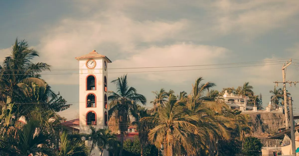 A picturesque beachside town with a prominent clock tower and lush palm trees under a sunny sky.