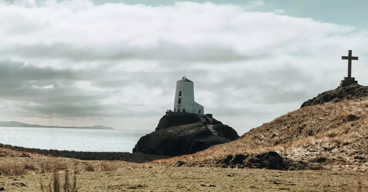 Scenic view of Twr Mawr Lighthouse and a cross on Ynys Llanddwyn Island with sea and cloudy sky.