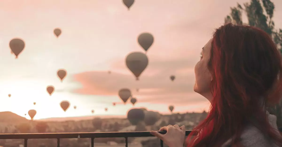 A woman with red hair watches hot air balloons soar over Cappadocia during a serene sunrise.