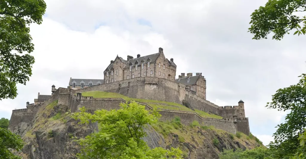 View of the iconic Edinburgh Castle perched on Castle Rock, framed by trees under a cloudy sky.