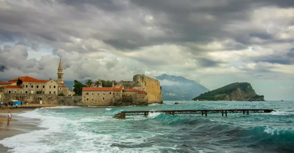 Historic Budva coastline with medieval architecture and rugged sea under dramatic skies.