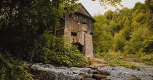 A rustic abandoned wooden mill surrounded by lush forest and a flowing stream under a cloudy sky.