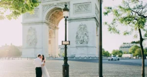 A couple in wedding attire embraces by the Arc de Triomphe in Paris on a sunny day.