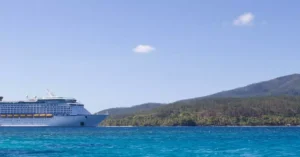 A large cruise ship positioned near a lush tropical island, set against a vibrant blue sky and ocean.