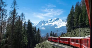 A red train travels through lush forests and snowy mountains under a clear blue sky in Switzerland.