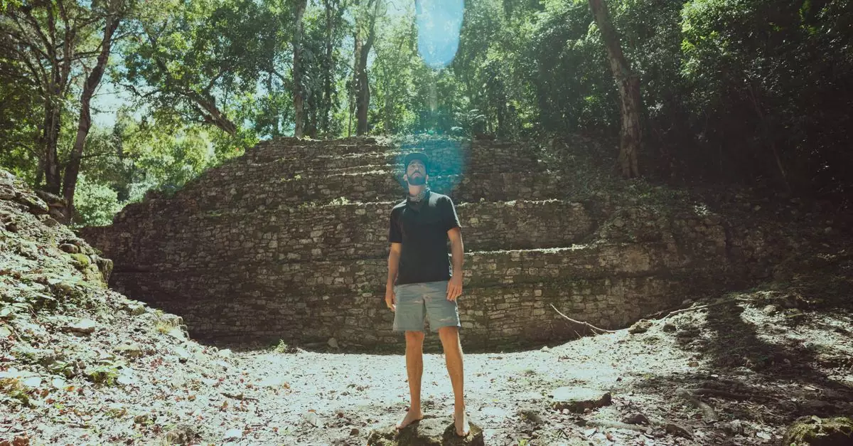 A man stands among ancient Mayan ruins, basking in sunlight filtering through the jungle canopy.