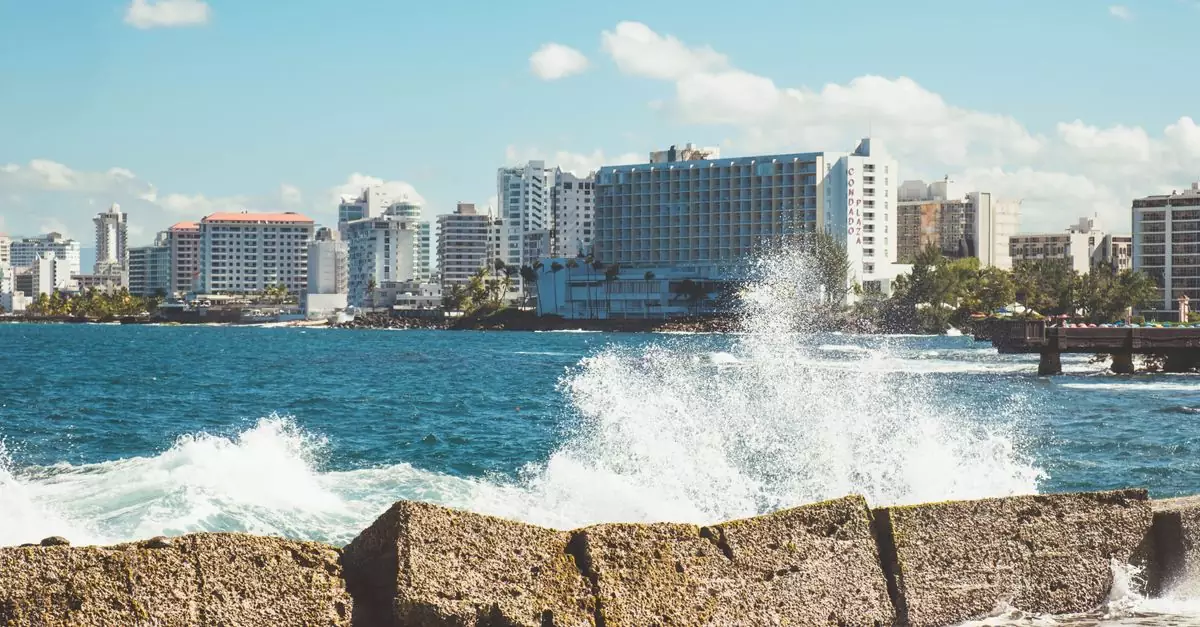 Wave crashing against a breakwater with San Juan's urban skyline in the background.