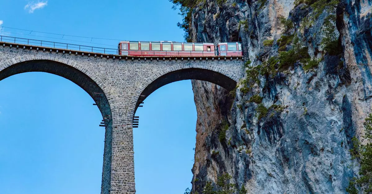 A train traverses the iconic Landwasser Viaduct against a blue sky and rugged cliffs in Switzerland.