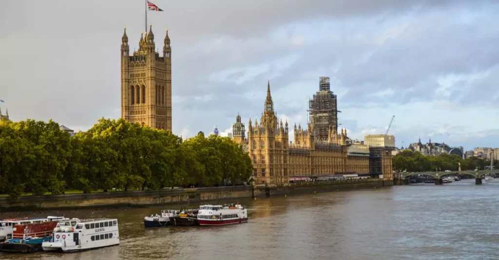 Best Royal Destinations England: Palaces & Castles 2 Exterior of aged building of the Houses of Parliament on bushy coast near river with vessels under cloudy sky of London