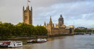 Exterior of aged building of the Houses of Parliament on bushy coast near river with vessels under cloudy sky of London