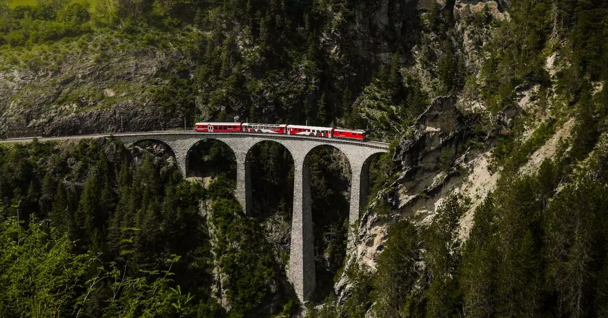 Red passenger train traveling on a stone viaduct, surrounded by lush green alpine scenery.