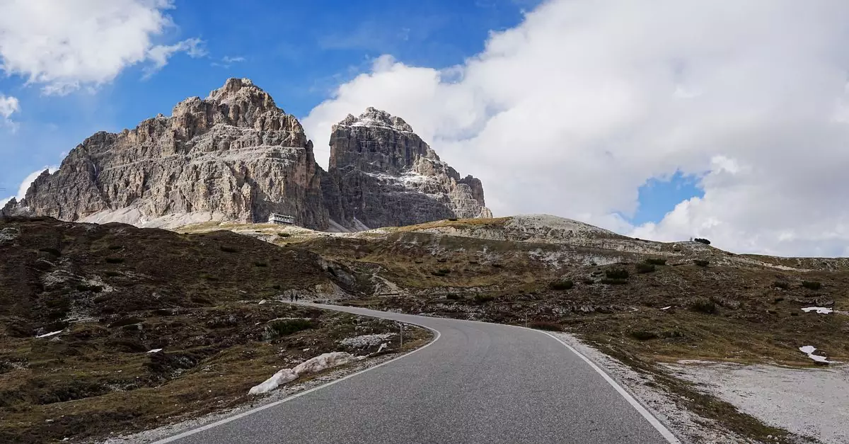 A winding road approaches the iconic Tre Cime di Lavaredo under a bright blue sky.