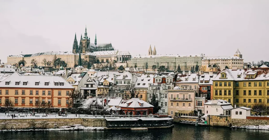 Explore a stunning winter view of Prague with snow-covered rooftops and historic architecture along the river.