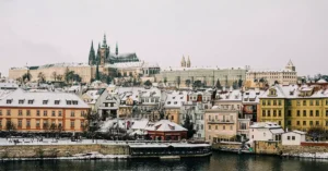 Explore a stunning winter view of Prague with snow-covered rooftops and historic architecture along the river.