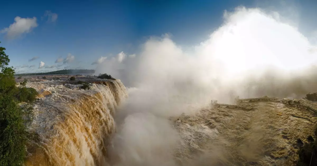 Panoramic view of the powerful Iguazu Falls surrounded by mist and lush greenery under a clear blue sky.