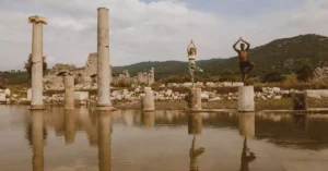 Two people practicing yoga poses on ancient stone ruins beside water. Reflective and serene atmosphere.