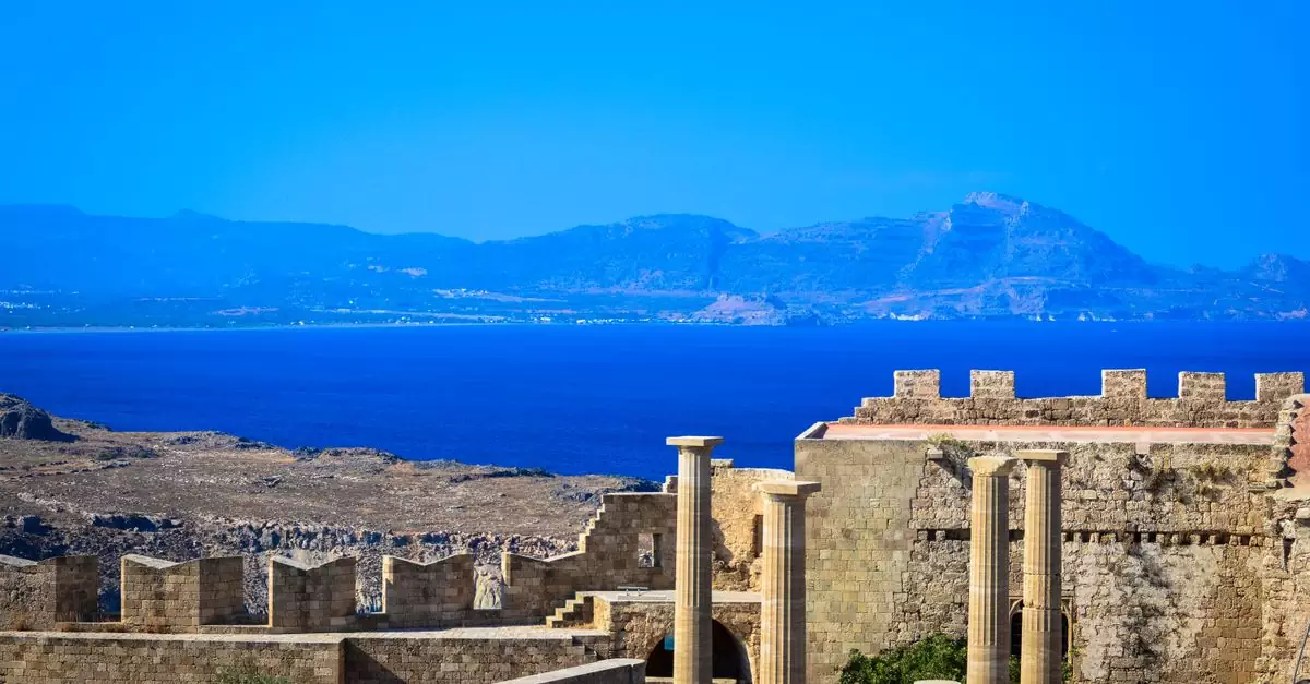 Scenic view of ancient Greek ruins with stone columns and walls against a vibrant blue Aegean Sea backdrop.