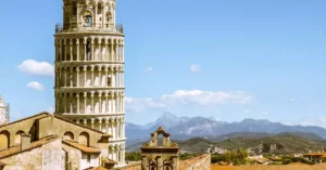 Leaning Tower of Pisa in Italy framed by rooftops and distant mountains under a clear blue sky.