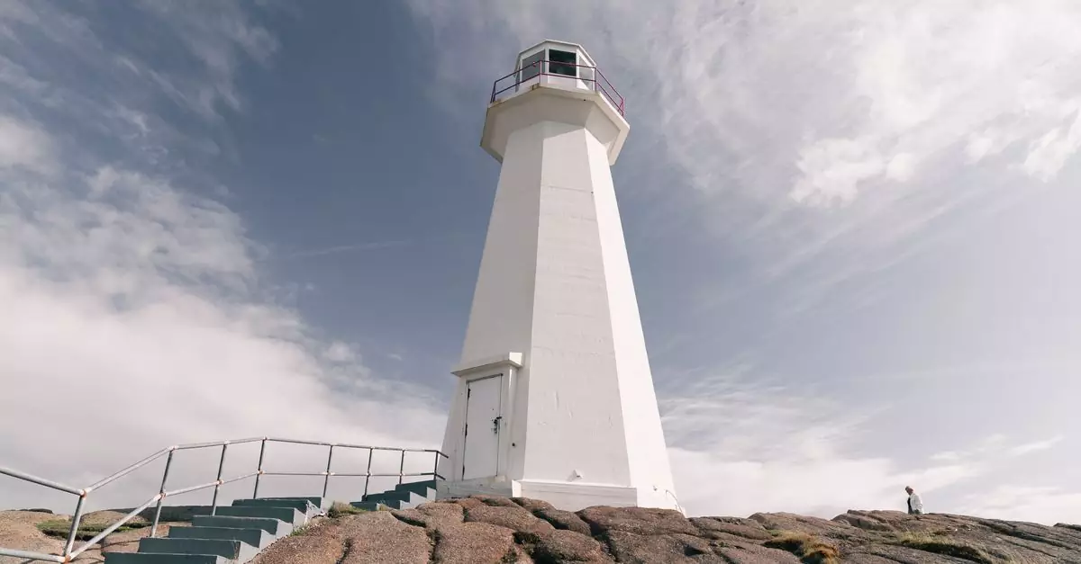 White lighthouse on rocky terrain with stairs, under a bright cloudy sky.