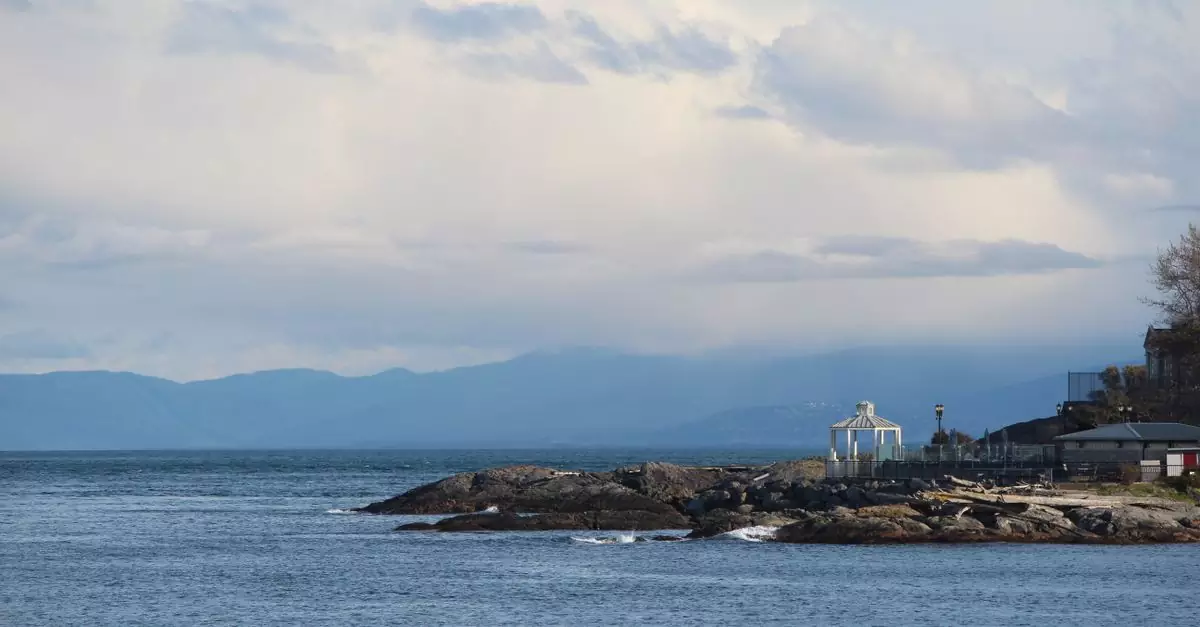 A beautiful coastal landscape of Victoria, BC depicting the rocky shoreline and distant mountains under a cloudy sky.